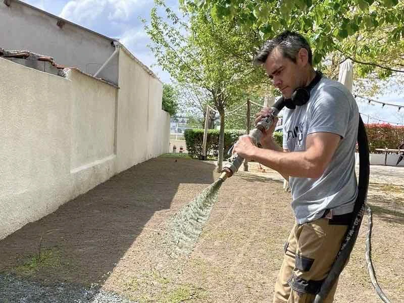 Un opérateur Rapid Gazon projette le mélange de graines et de mulch sur la terre nue d'un jardin de particulier, le long d'un mur blanc