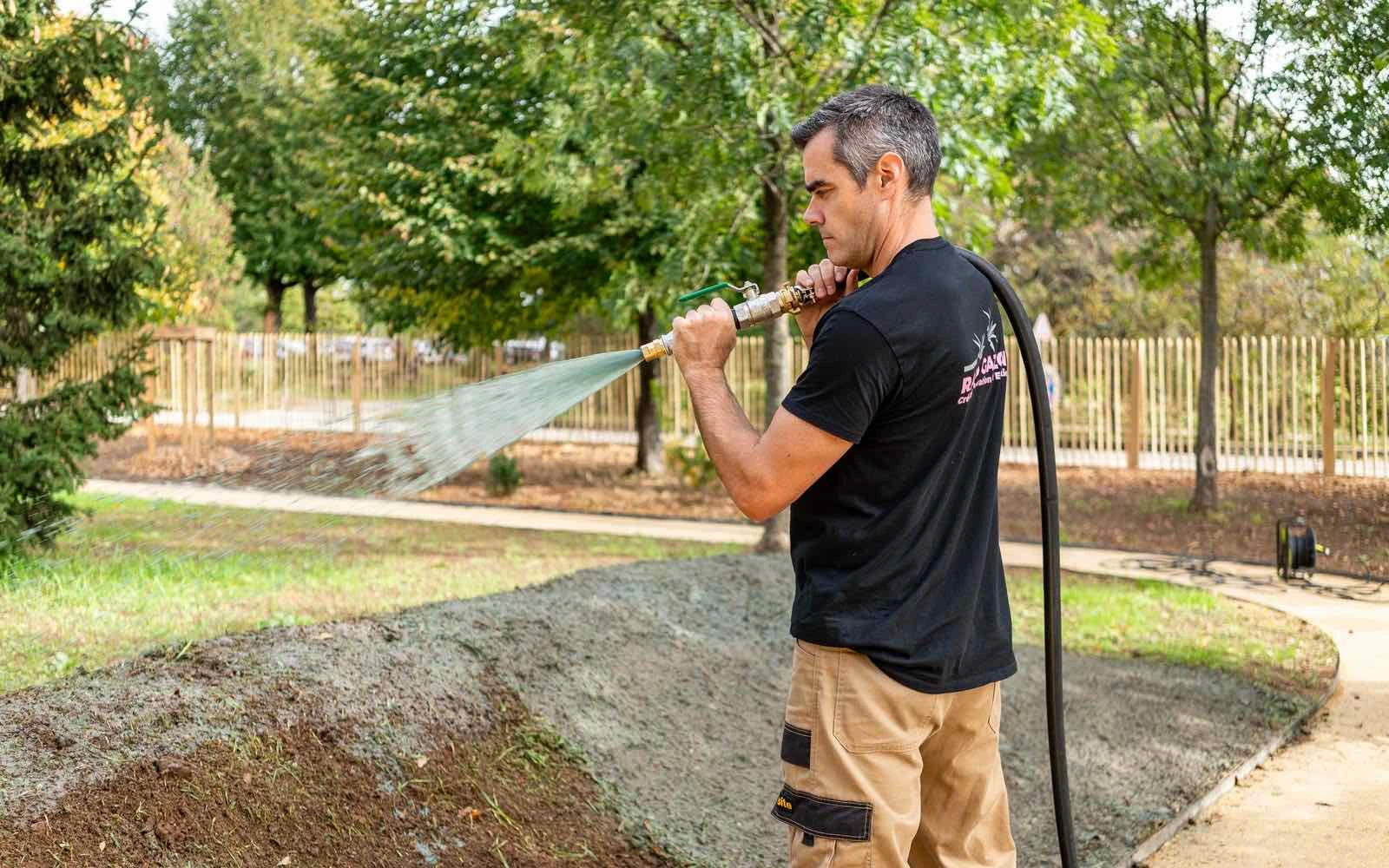 Un opérateur en t-shirt noir Rapid Gazon projette à la lance un mélange gris-vert sur une butte de terre dans un parc arboré